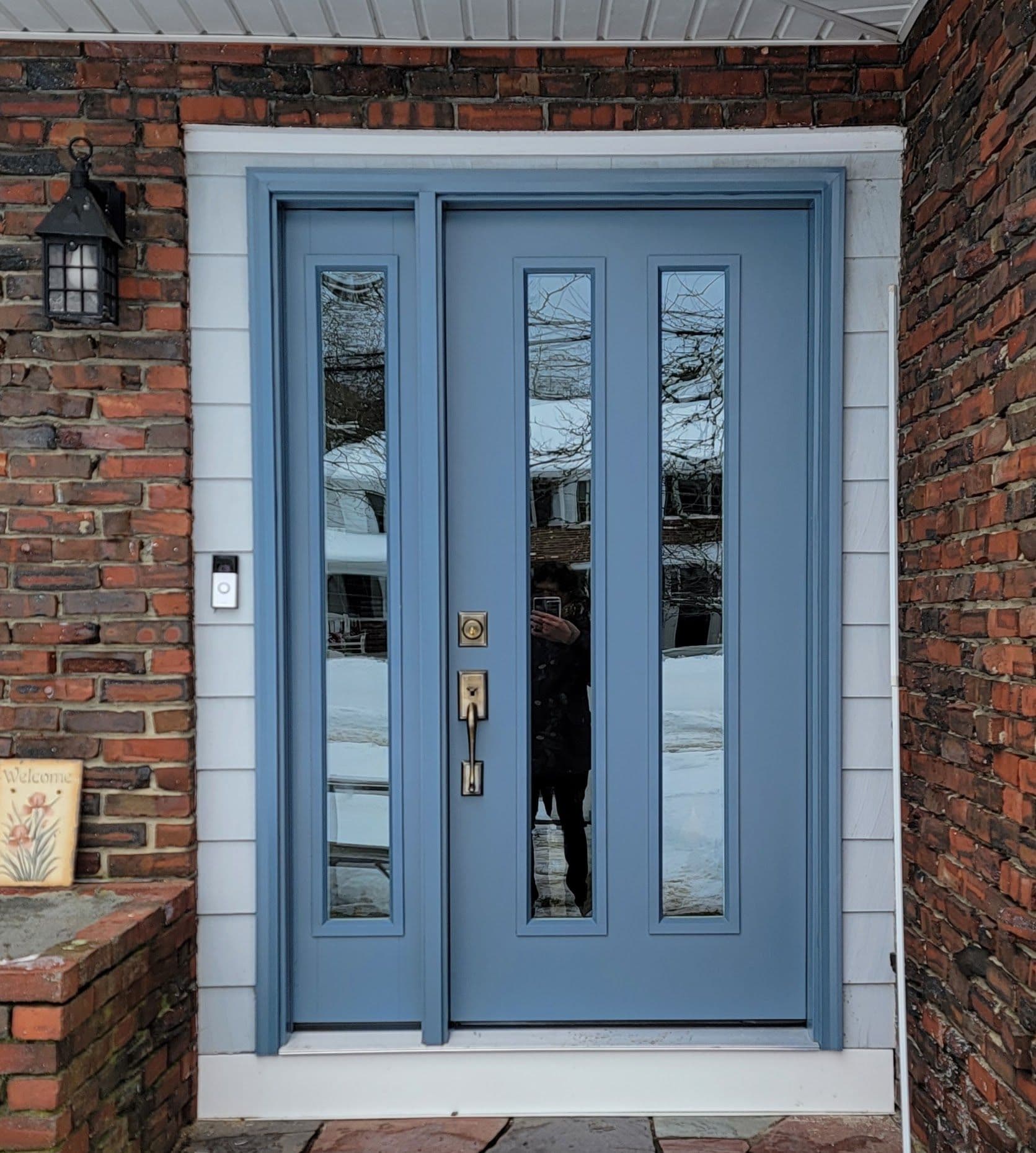 A blue front door set in a brick facade with vertical glass panels, reflecting a snowy winter scene. Side light and a small "Welcome" sign add warmth.