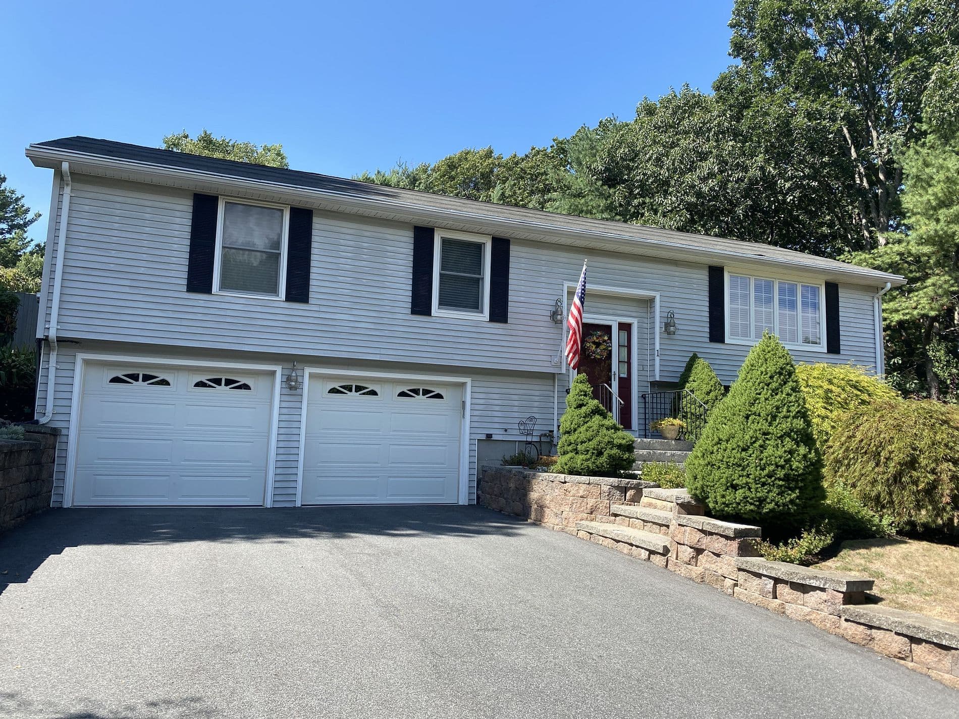 A suburban split-level house with white siding and black shutters. A U.S. flag is near the door, with green shrubs and stone steps leading up. Bright, clear day.