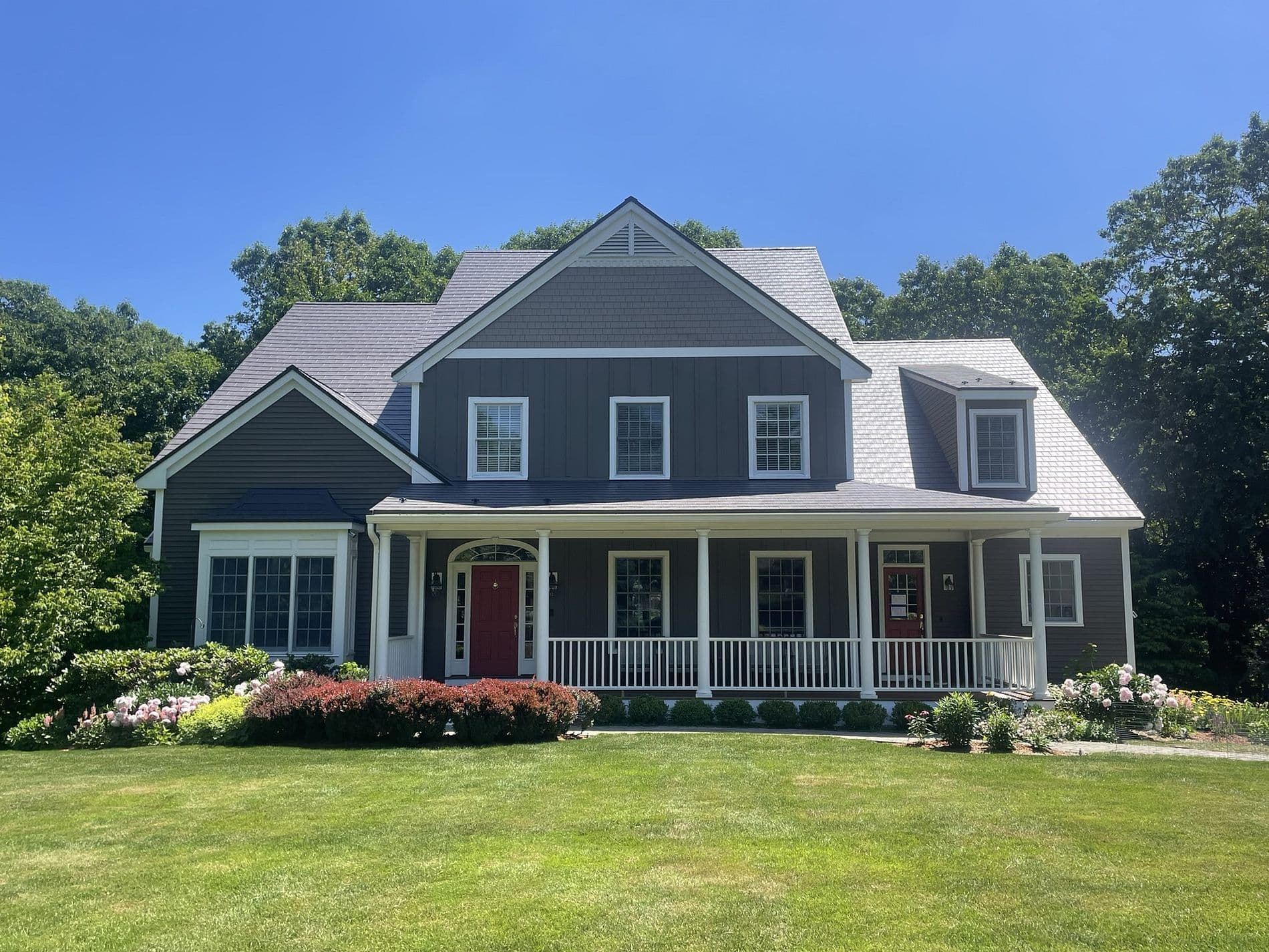 A spacious, two-story gray house with a large front porch and red doors, surrounded by a manicured lawn, colorful flowerbeds, and lush trees under a clear blue sky.
