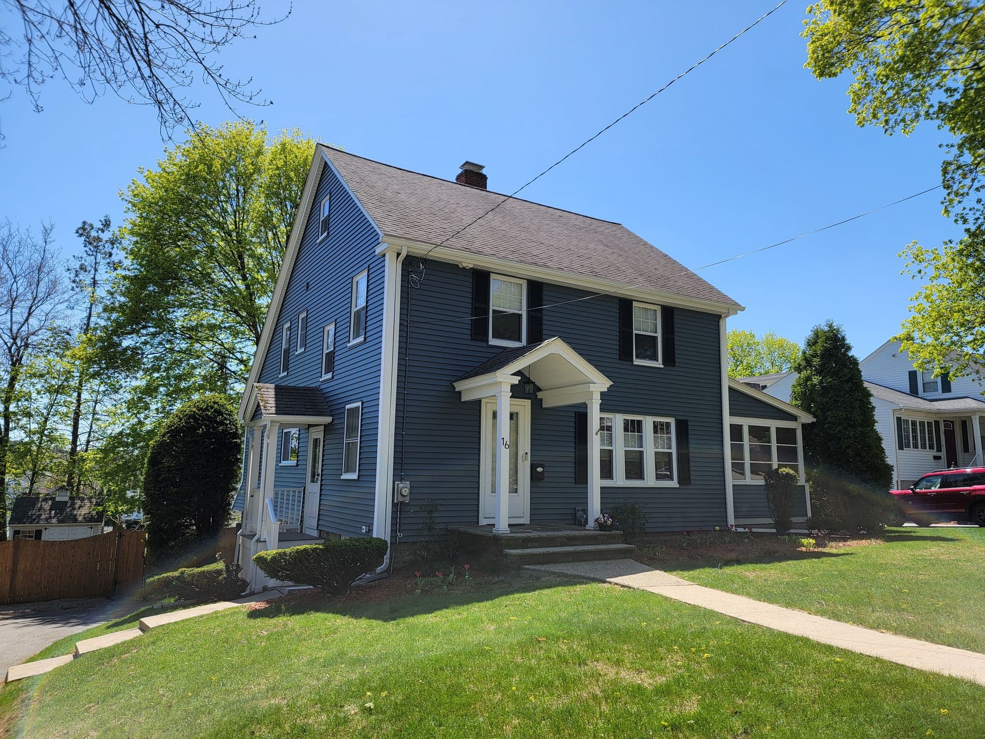 A charming blue two-story house with white trim and black shutters. It sits on a lush green lawn with a clear blue sky and trees in the background.