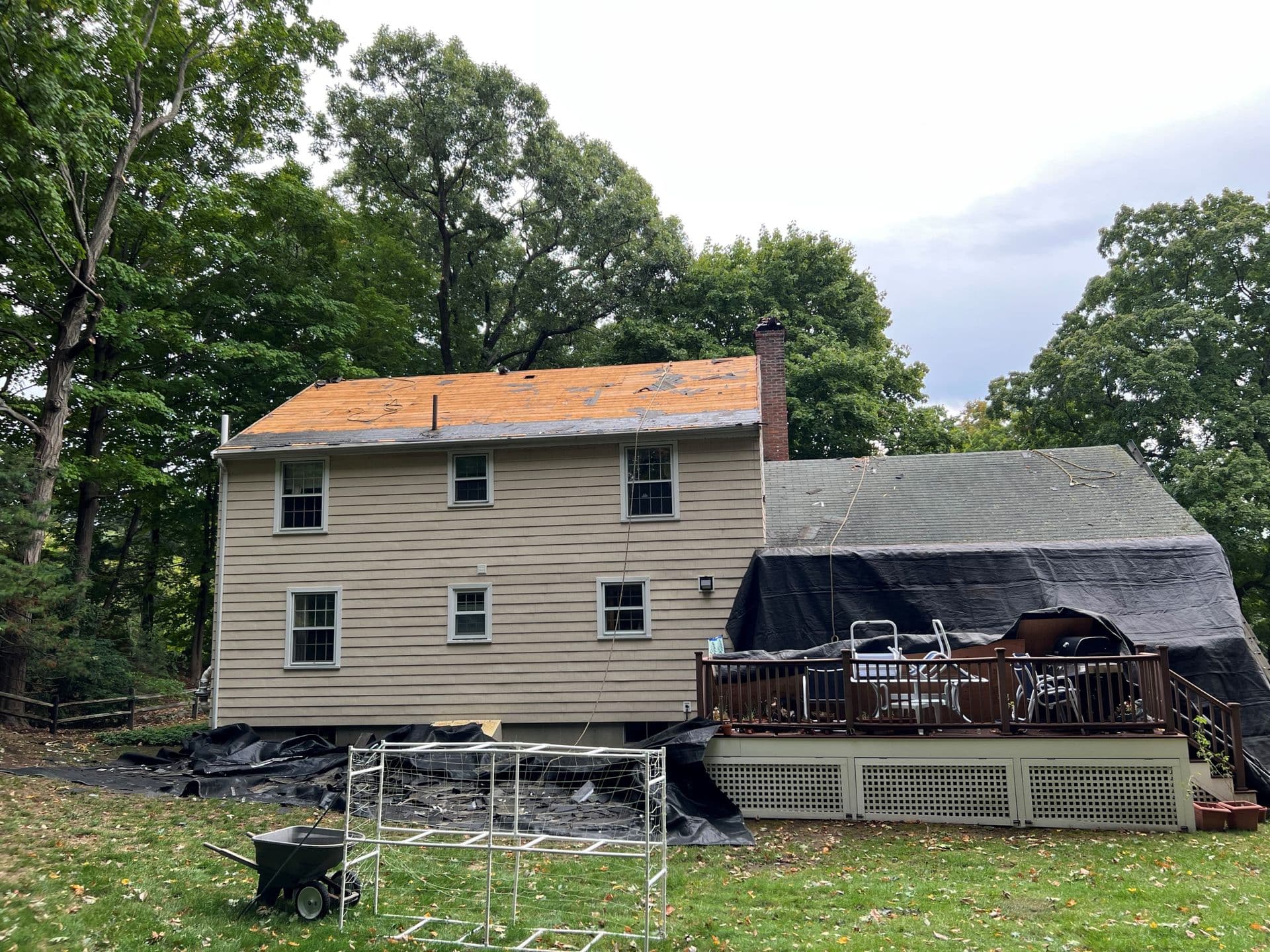 A beige two-story house is undergoing roof repairs, with tarps and protective materials covering sections. A wooden deck holds patio furniture. Lush trees surround the yard, and a wheelbarrow is in the foreground, hinting at ongoing work. The scene conveys a busy, work-in-progress atmosphere.