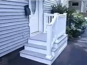 A white exterior door with three stairs and a white railing leads to a small porch on a gray-sided house. Plants and a walkway are visible nearby.