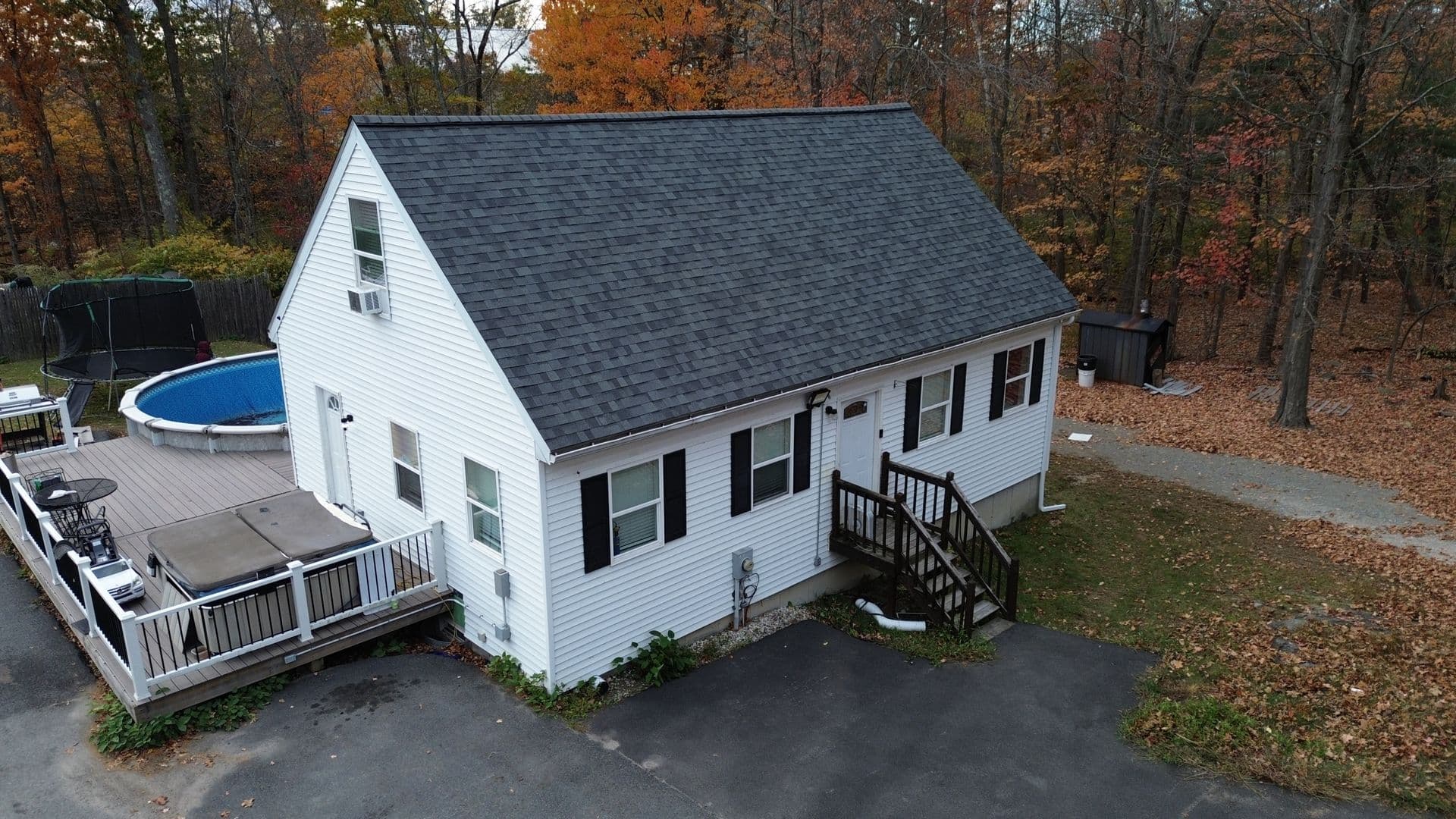 A white house with a dark roof sits surrounded by autumn trees. A deck with a jacuzzi and round pool is on the left, creating a cozy outdoor setting.
