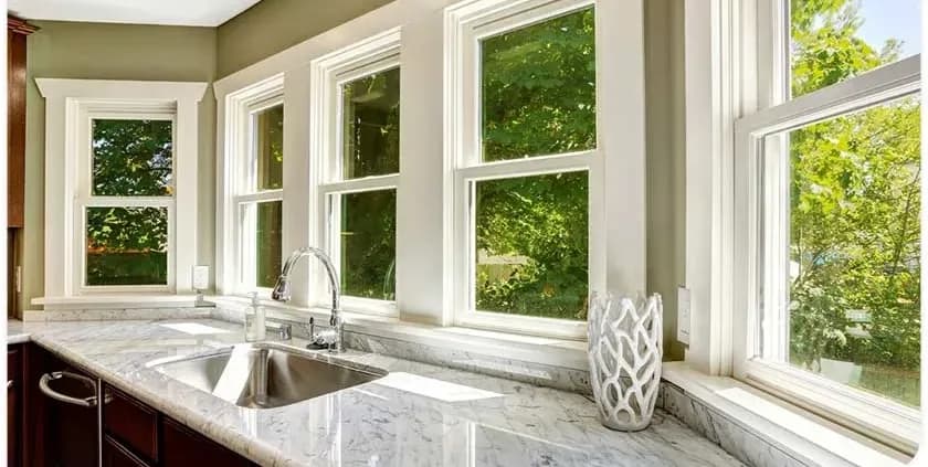 Bright kitchen with large windows, white trim, and lush green views. Marble countertop features a silver sink and decorative vase, creating a serene atmosphere.