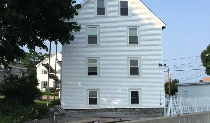 A tall, narrow white house with six windows is seen against a clear blue sky. It's surrounded by trees and adjacent buildings, creating a calm suburban scene.