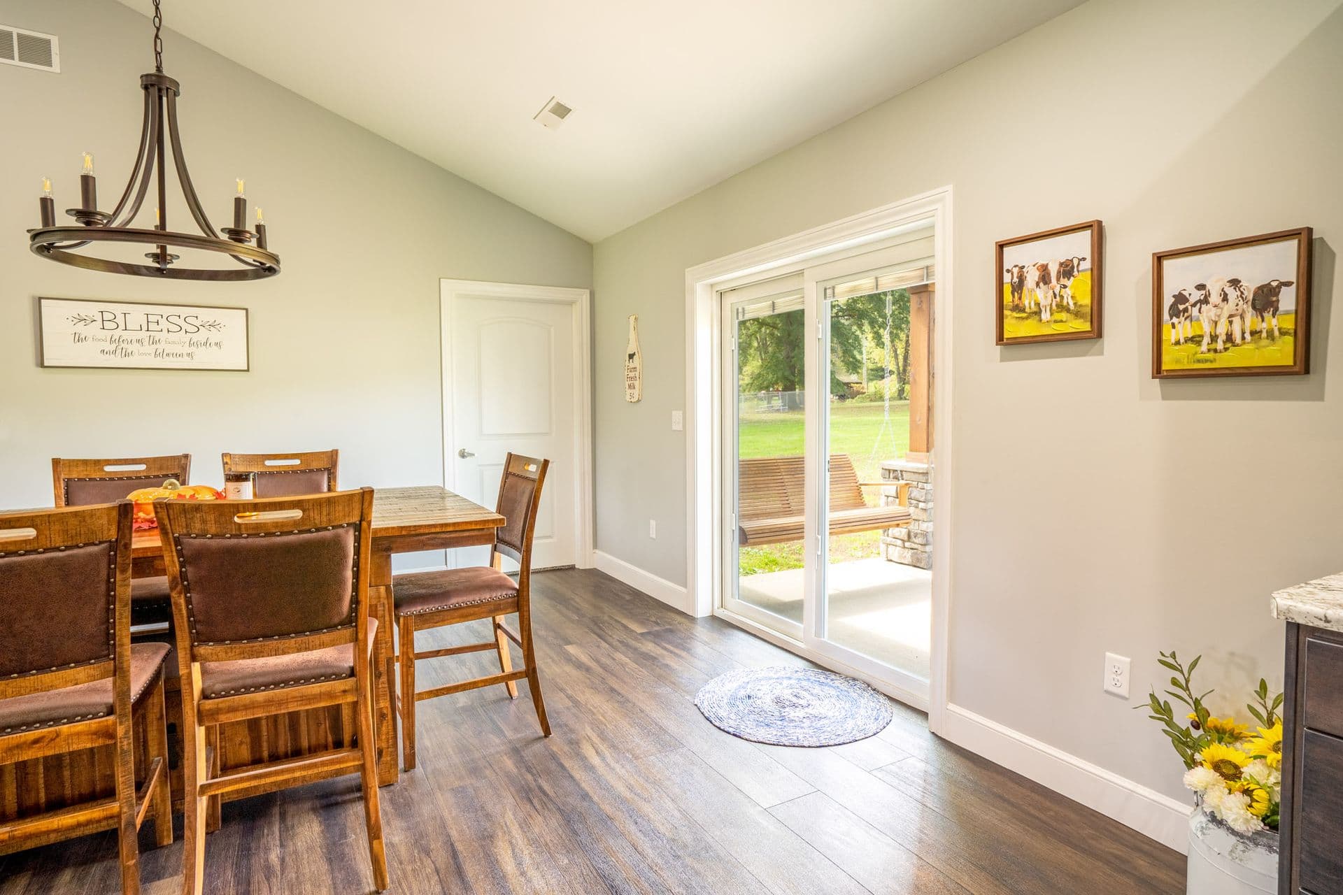 Dining room with wooden table and six chairs, a chandelier, and a sign on the wall. Glass doors open to a patio. Bright, airy ambiance.