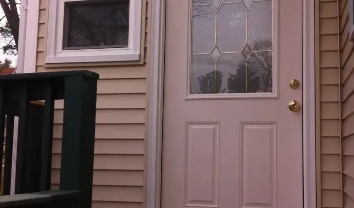 A beige door with decorative glass panels is set in a house with light siding. A small window is on the left, and a dark green railing is partially visible.