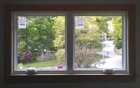 A large window view of a lush, sunny garden with blooming purple flowers and a winding road. Green trees and a peaceful suburban scene are visible.