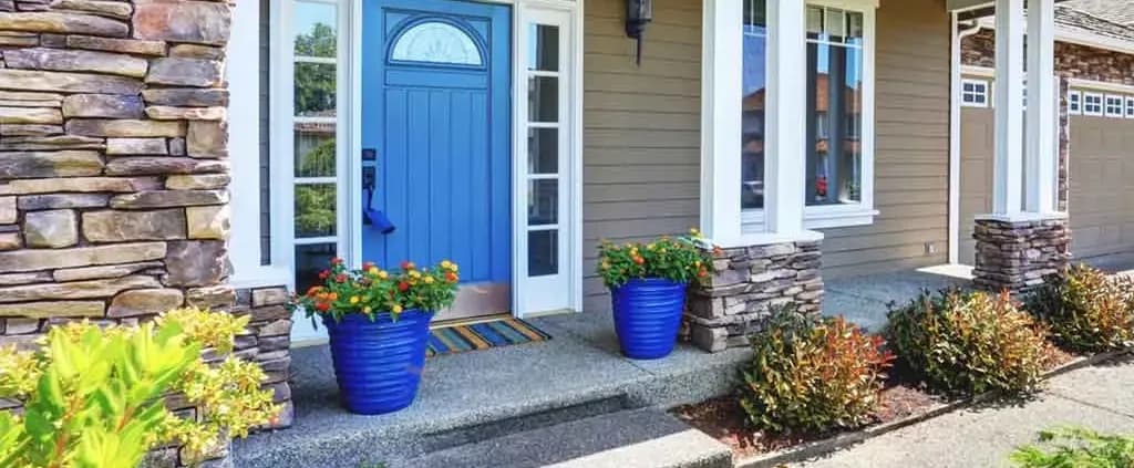 Front porch of a house with a bright blue door framed by sidelights, flanked by two large blue flower pots with vibrant orange flowers, on a stone-lined pathway.