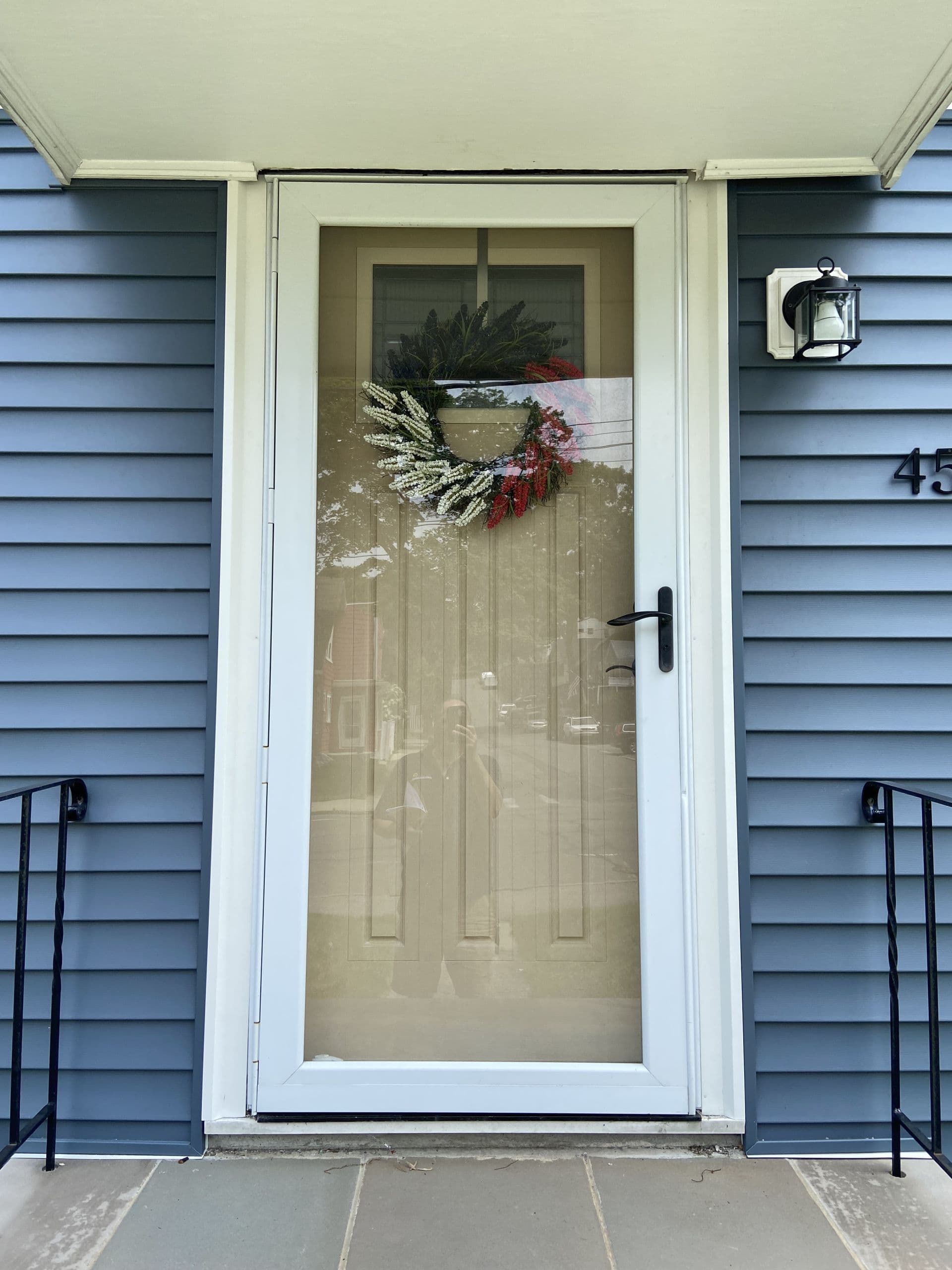 Blue-sided house with a white door featuring a decorative wreath of green, white, and red leaves. A sleek black handle and lamp are on the right.
