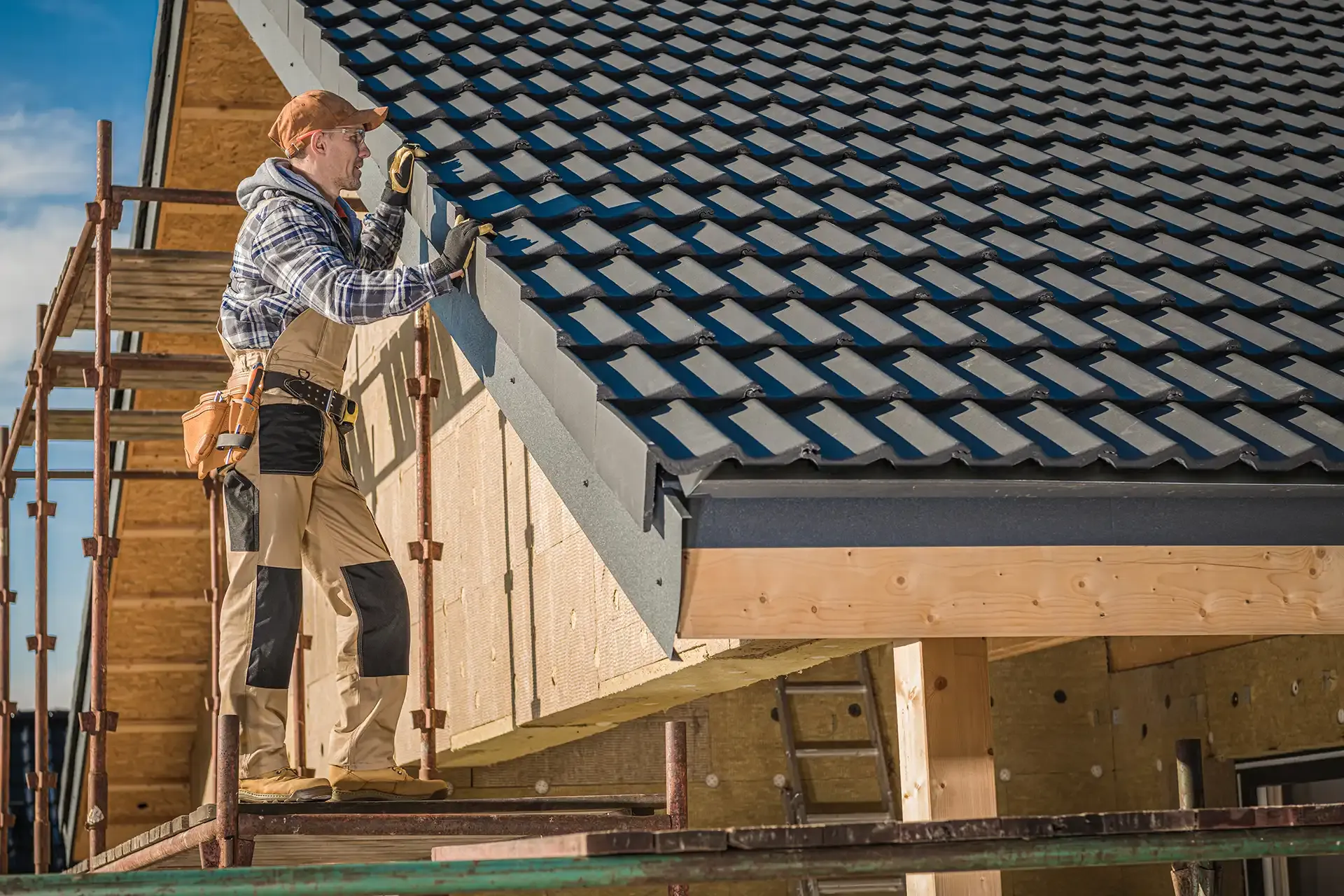 A construction worker in safety gear installs a dark metal roof on a wooden house frame, standing on scaffolding under a clear blue sky, focused and diligent.
