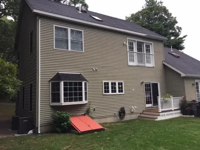 Two-story beige house with dark roof, double windows, small balcony, and red cellar doors. A wooden deck steps down to a grassy yard, surrounded by trees.