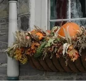 A window box display features a pumpkin surrounded by autumn leaves, dried grasses, and small gourds against a stone wall, evoking a cozy, fall ambiance.