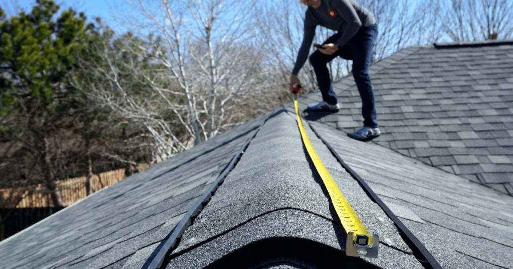 A person is on a roof, measuring the ridge with a yellow tape measure. The shingles are dark gray, and trees are visible in the background under a clear sky.