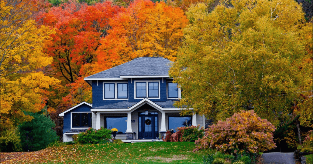 A two-story blue house is surrounded by vibrant autumn foliage in shades of orange, yellow, and red, creating a cozy and picturesque fall scene.