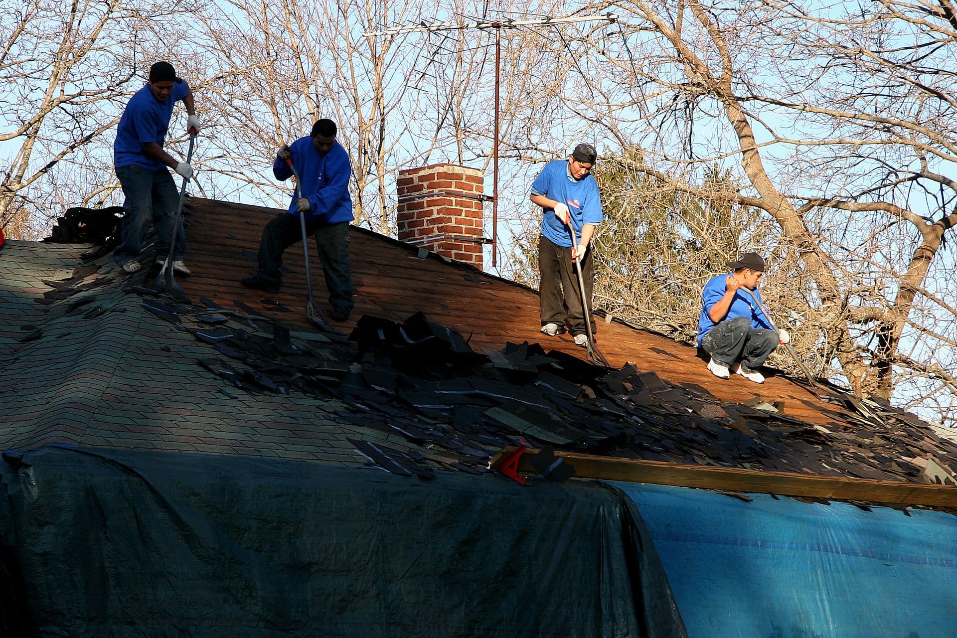Four workers in blue shirts remove shingles from a roof on a sunny day. Bare trees and a chimney in the background suggest a winter setting.