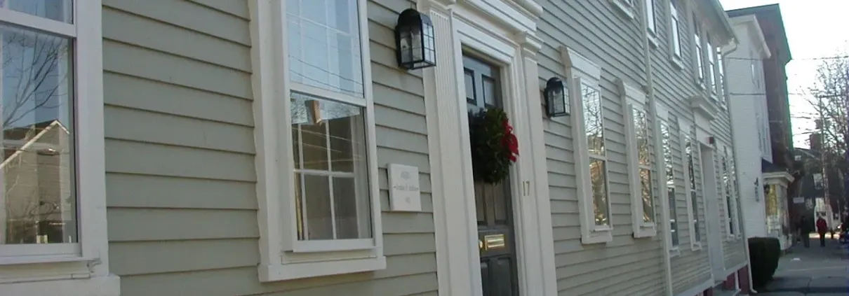 A historic-style row house with light gray siding and white trim. The entrance features a black door adorned with a festive wreath, creating a welcoming atmosphere.