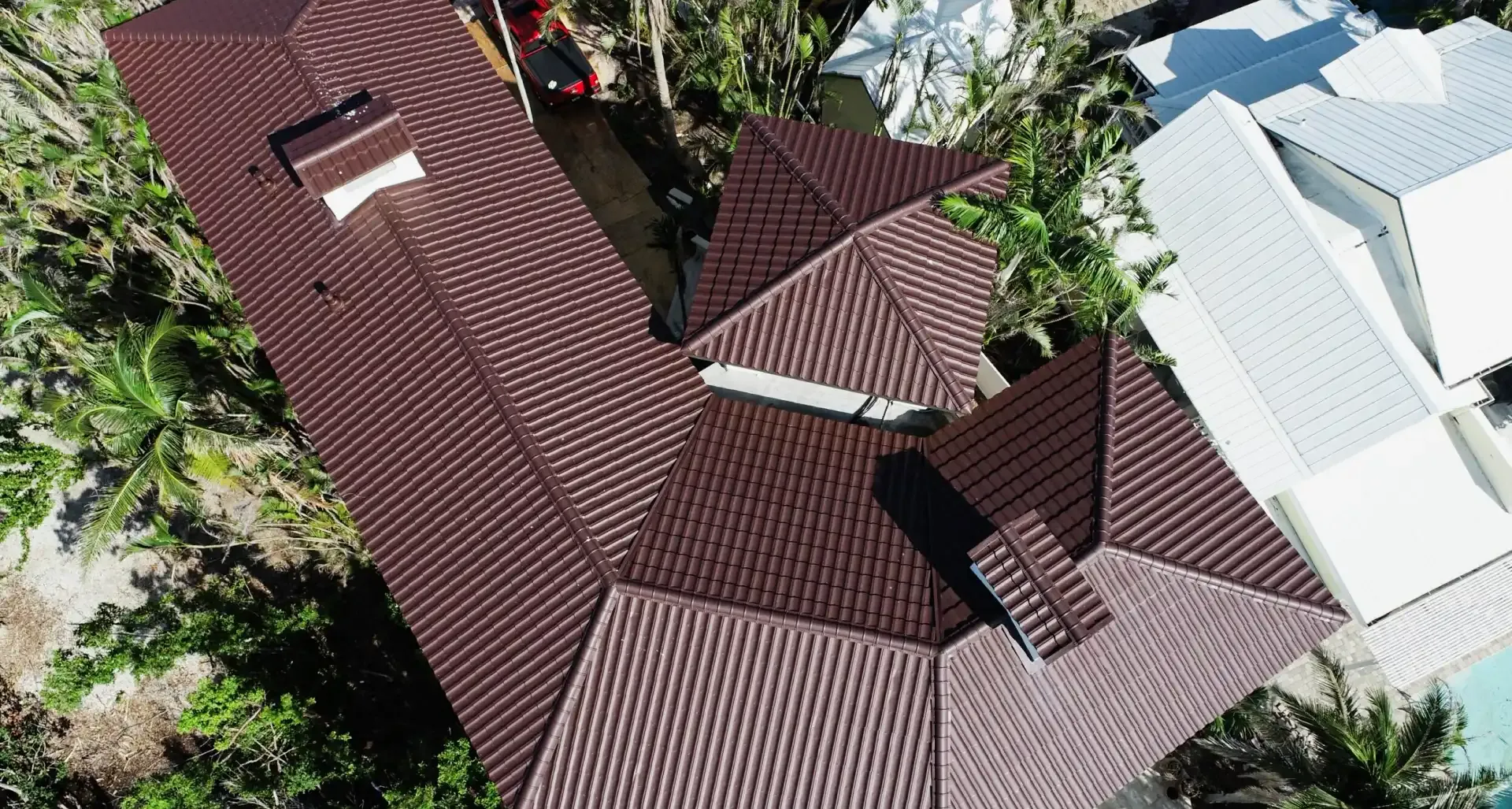 Aerial view of a house with complex, brown-tiled roofing contrasted against a neighboring white-roofed building. Surrounding lush greenery adds texture.