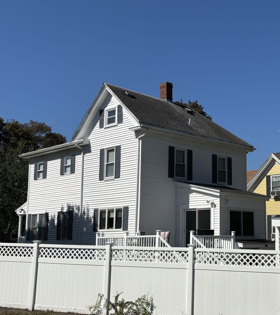 A white two-story house with dark shutters and a chimney sits behind a white lattice fence. The sky is clear blue, suggesting a bright, sunny day.