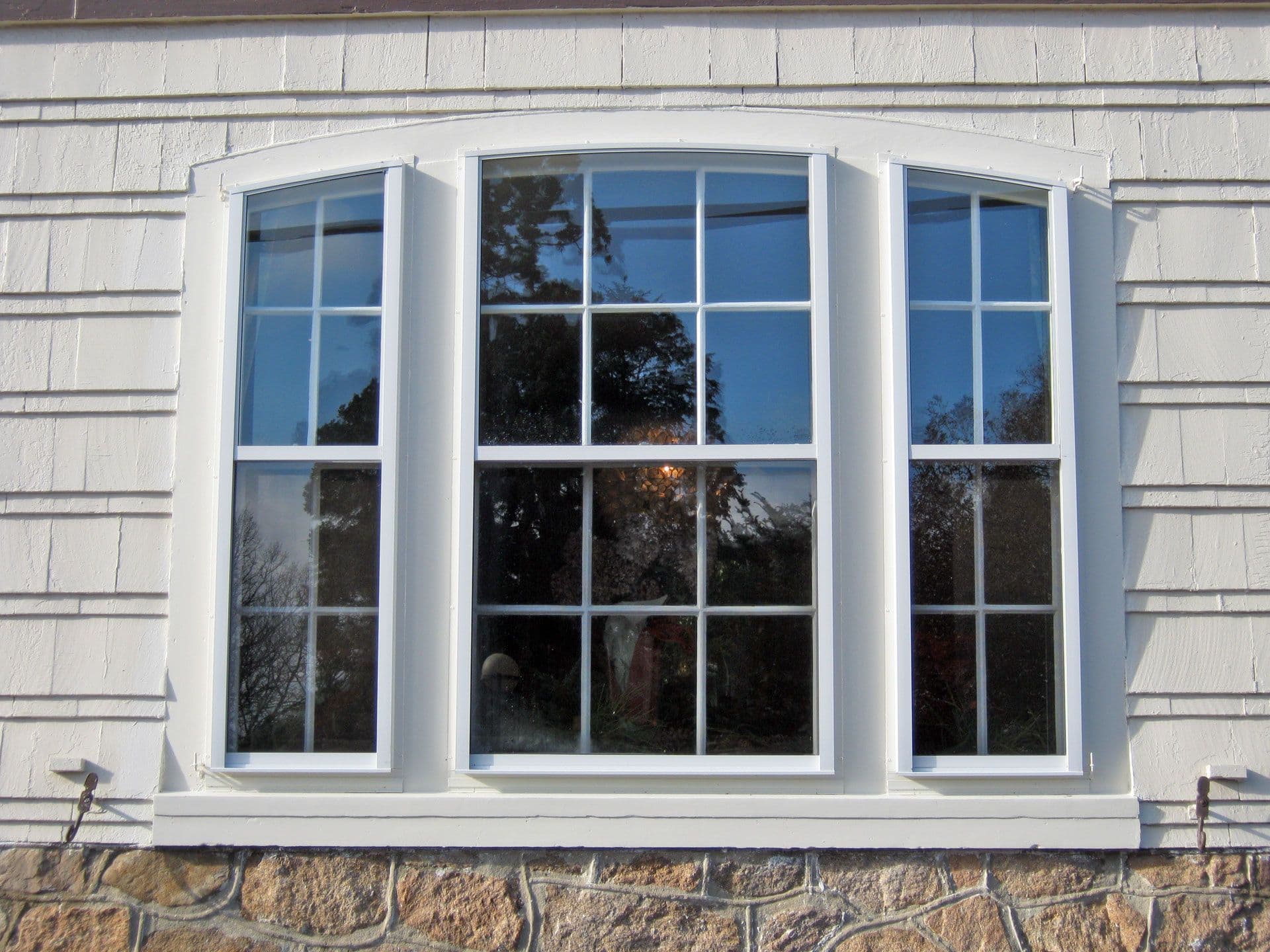 Arched white-framed window with three panels, each having multiple panes, set in a light-colored wooden wall above a stone foundation.