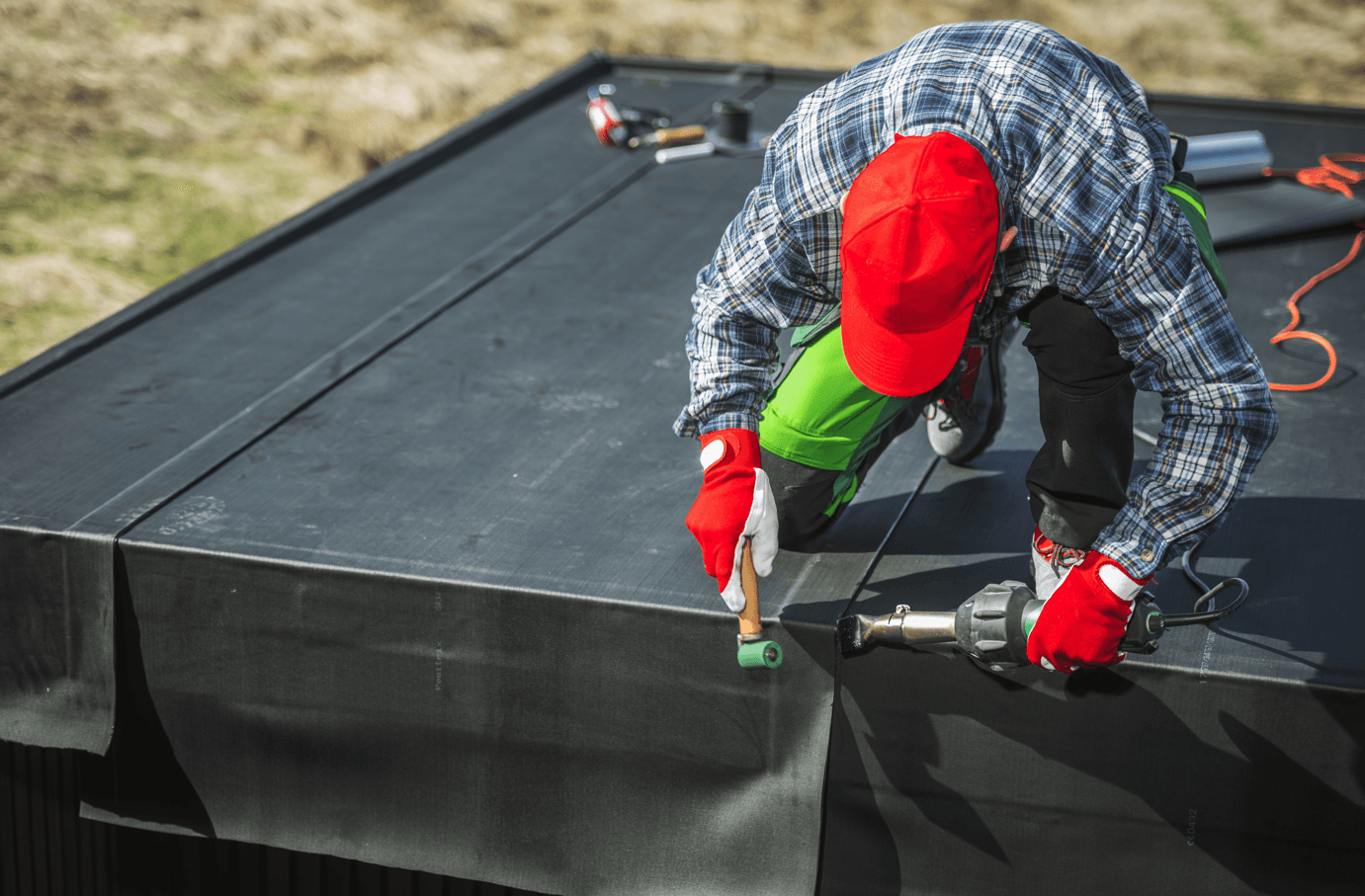 A worker in red gloves and a hat installs a black rubber roof, using tools on a flat rooftop under natural light, focusing intently on the task.