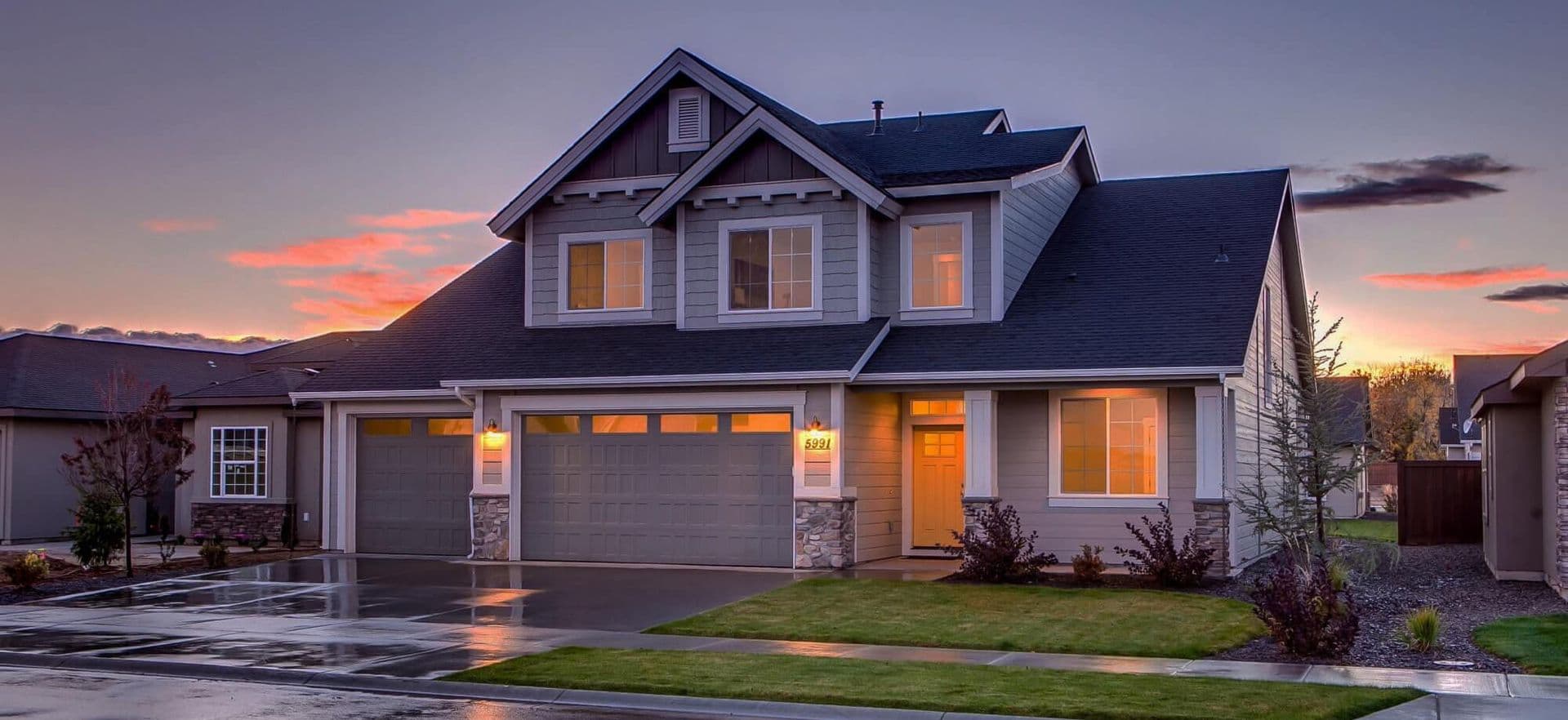 A two-story suburban house at dusk, with lit windows, wet driveway, and a manicured lawn. The sky is colorful with pink and orange hues.
