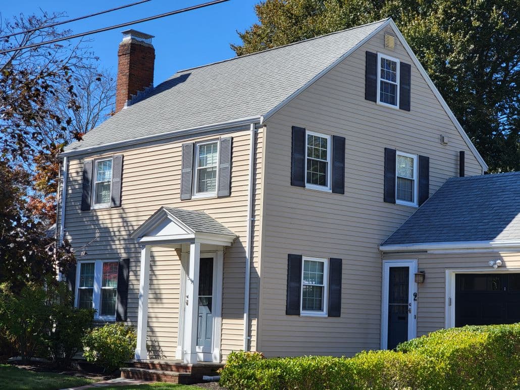 Two-story beige house with gray shutters and a gabled roof, featuring a white front door and chimney. Surrounded by shrubs and trees under a clear sky.