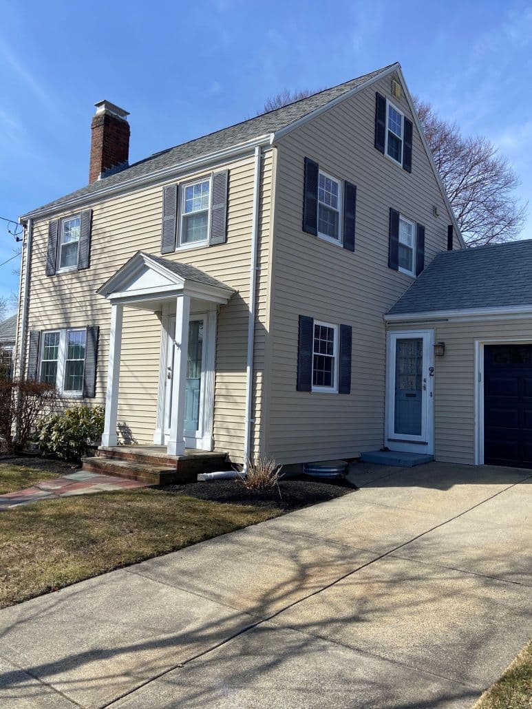 Two-story beige house with blue shutters and white trim, featuring a brick chimney and a well-maintained lawn. A sunny and clear day.