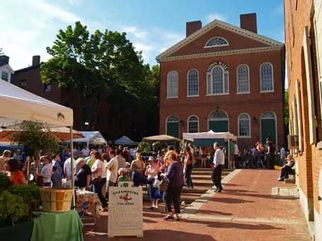 A lively outdoor market in front of a historic brick building with large arched windows. People browse stalls, enjoying a sunny day with trees nearby.
