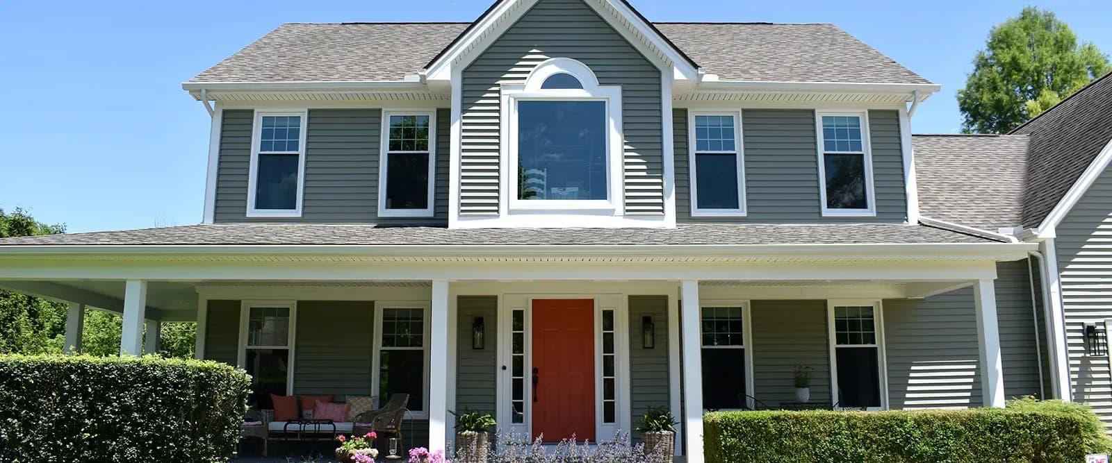 Two-story house with gray siding, symmetrical windows, red front door, and wraparound porch. Surrounded by bushes, under a clear blue sky. Inviting and peaceful.
