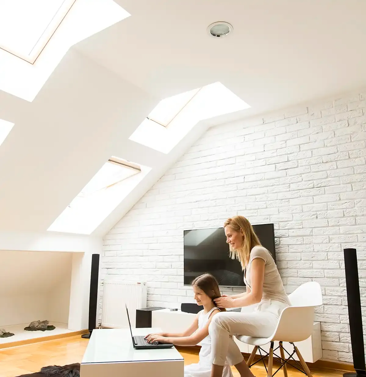 A woman kneels beside a child at a laptop in a bright, modern loft with skylights and white brick walls. They appear happy and engaged.