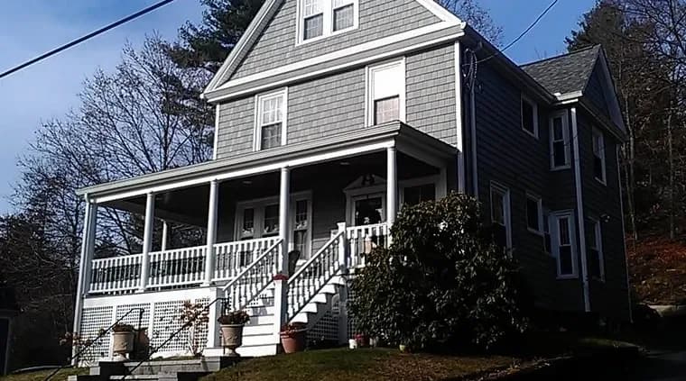 Gray, two-story house with white trim and a welcoming porch. The yard is neatly landscaped with shrubs. The scene is calm and sunlit.
