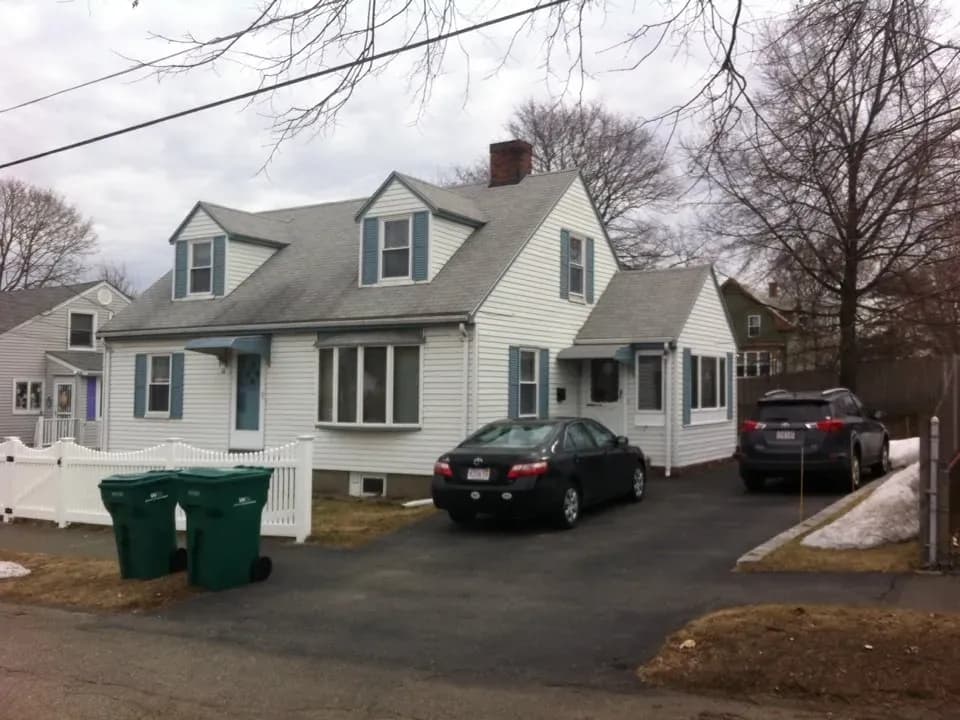 A cozy, white, two-story house with a pitched roof and chimney, flanked by two cars in the driveway. Leafless trees and a partly snowy yard create a wintry atmosphere.