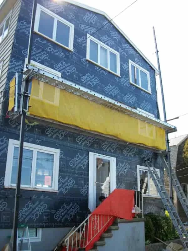 Three-story house under renovation with blue weatherproof wrap and yellow scaffolding mesh. A red cover protects the stairs. Tools are visible.