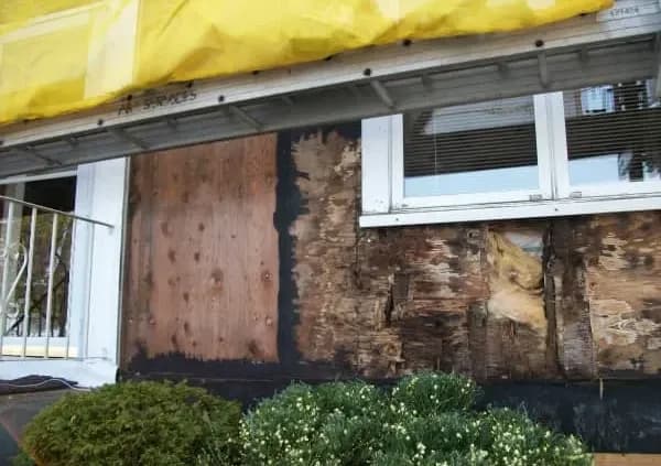 Exterior wall under repair, showing exposed plywood and decayed wood beneath a window. Yellow tarp covers a section above. Shrubs in the foreground.