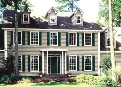A charming two-story house featuring a beige façade, green shutters, and multiple windows. Surrounded by trees, it conveys a welcoming, serene atmosphere.