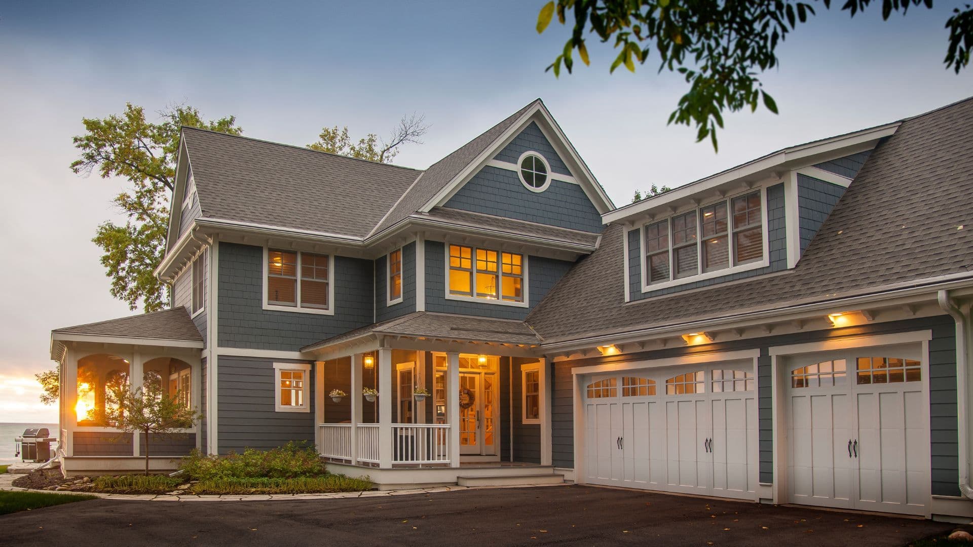 A charming two-story blue house with warm lit windows, a welcoming porch, and a double garage, surrounded by trees, at sunset, creating a cozy ambiance.