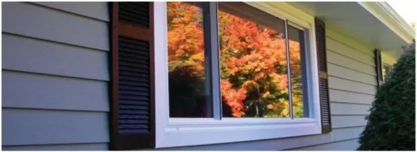 A window on a house exterior reflects vibrant autumn trees with orange and red leaves, framed by dark shutters. It creates a warm and serene autumnal ambiance.